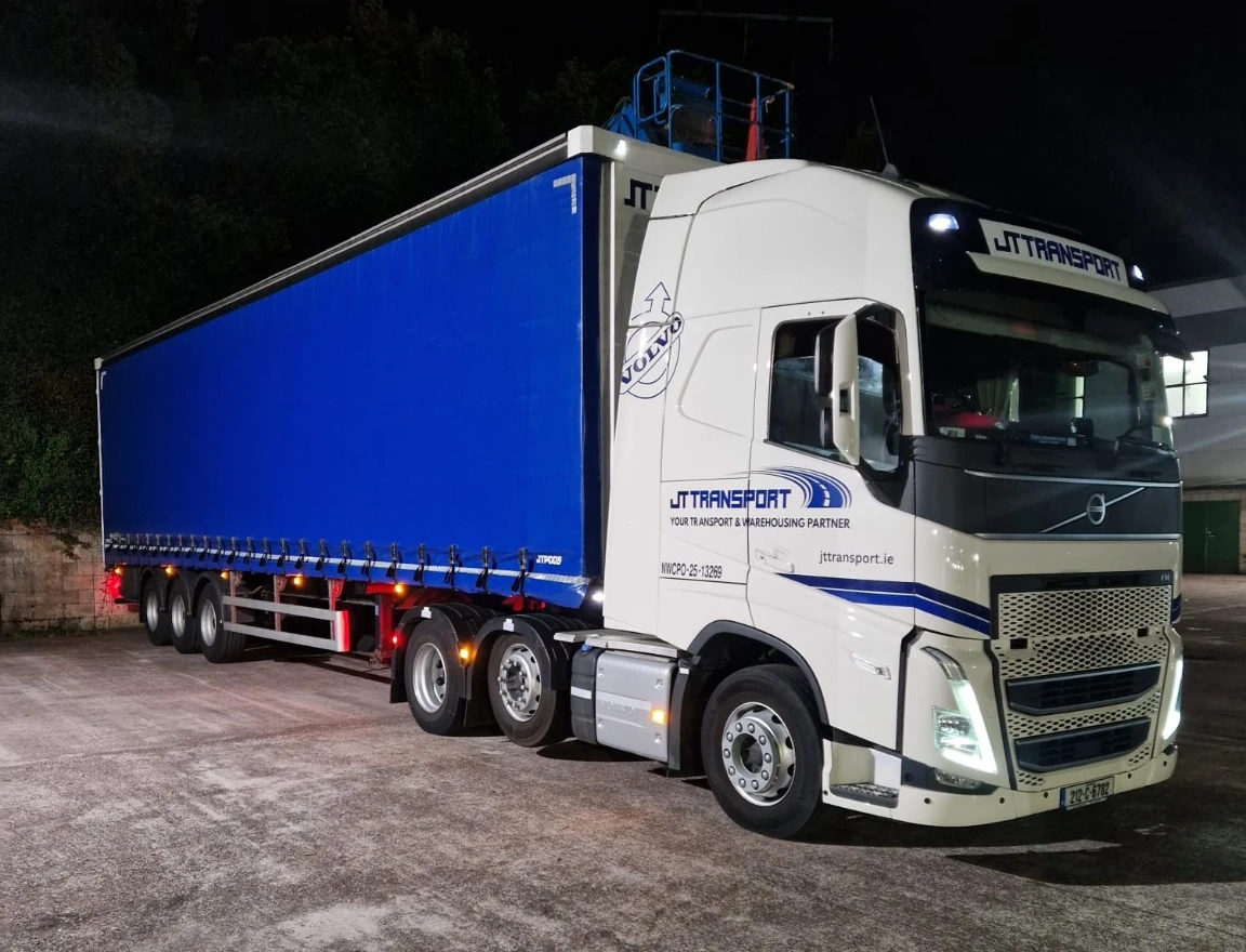 JT-branded Volvo articulated truck with a blue curtainsider trailer photographed at night.